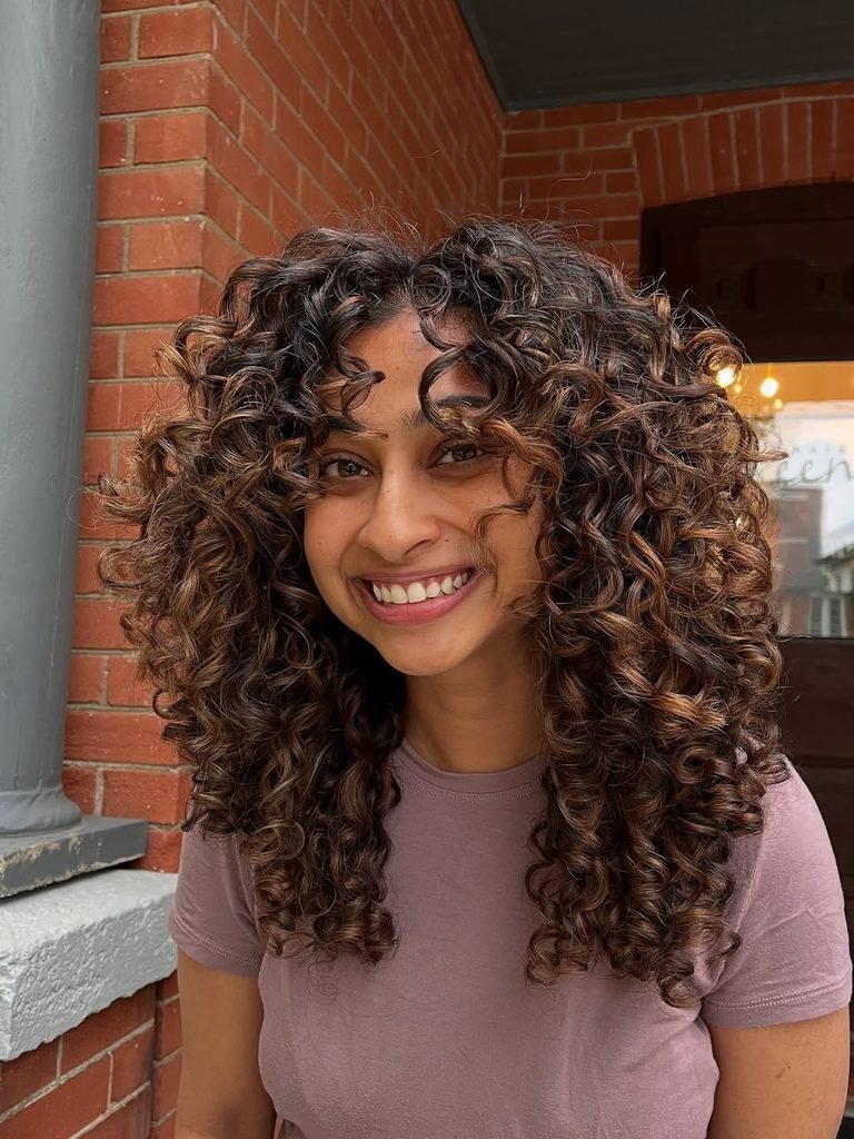 smiling woman with voluminous bouncy brunette curly hair and subtle caramel highlights