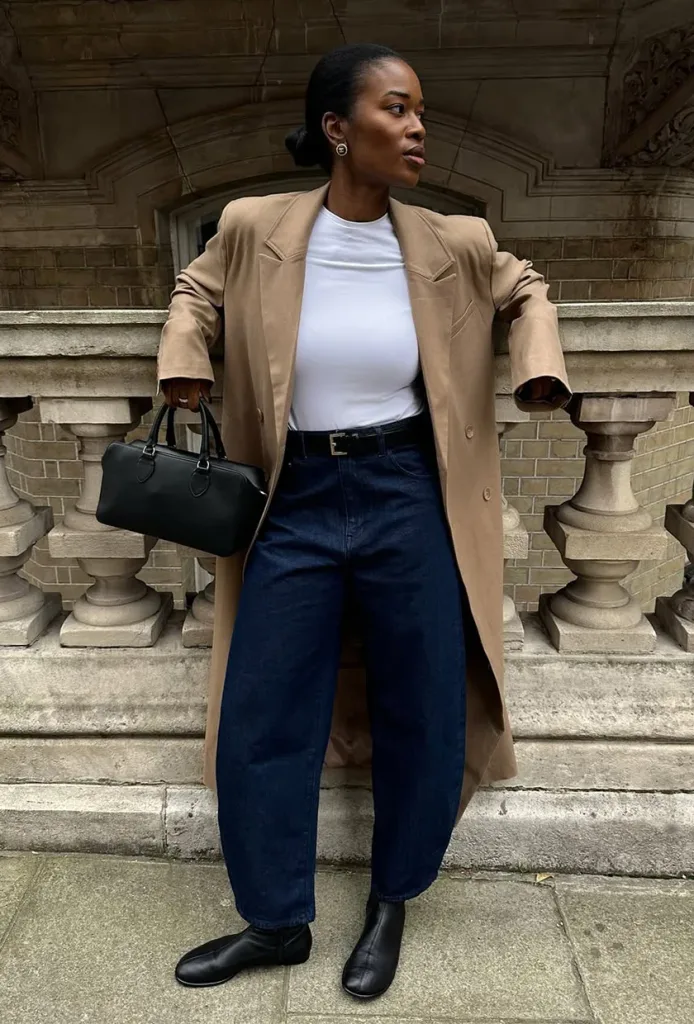 woman in tan trench coat and dark blue barrel jeans leaning on stone railing