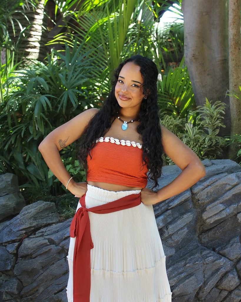 woman in an orange top and white skirt posing against lush green foliage