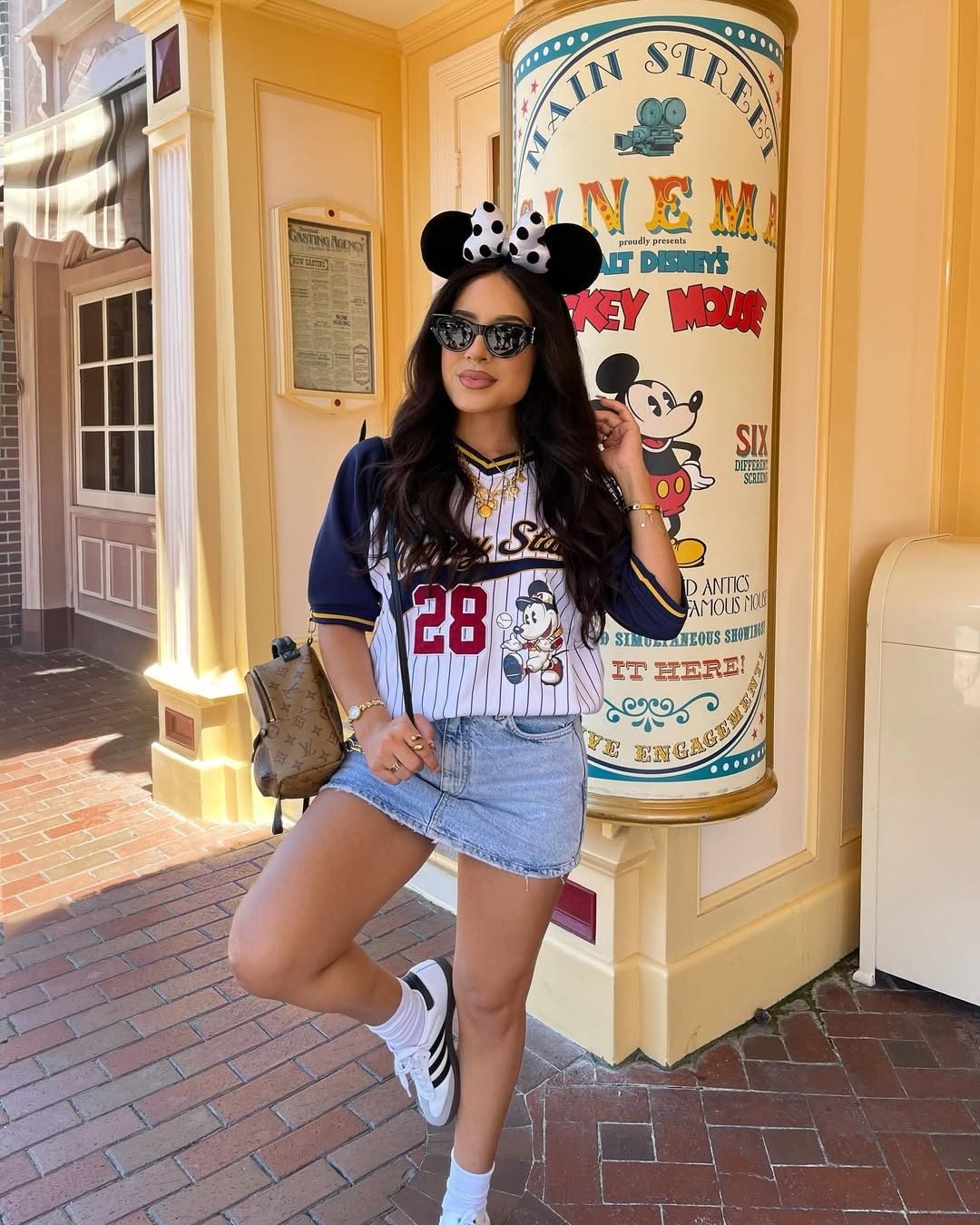 woman posing in minnie mouse ears, baseball jersey, and denim skirt at a disney park