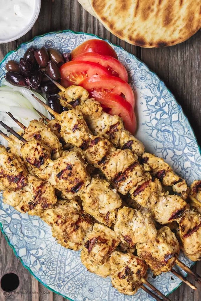 greek chicken souvlaki skewers on a blue platter with sliced tomatoes, olives, and pita bread