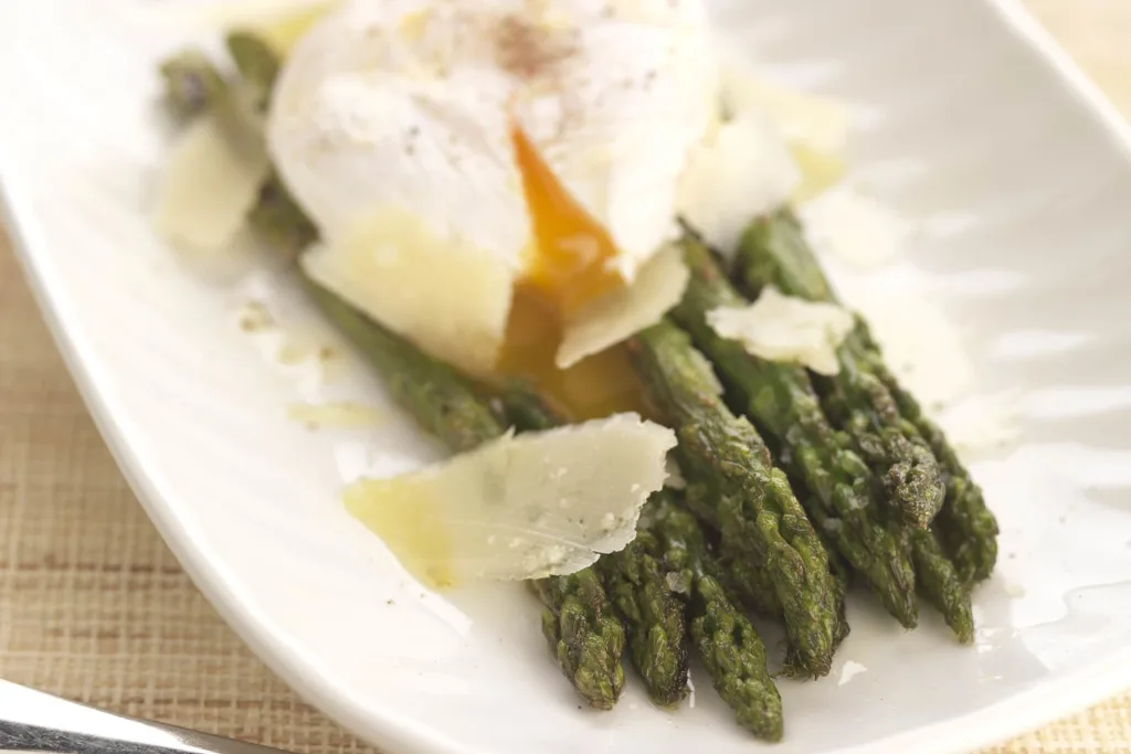 grilled asparagus spears on a white plate topped with a broken poached egg and shaved parmesan