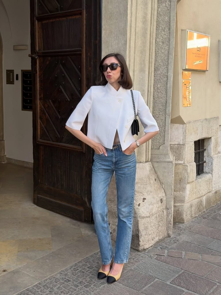 woman wearing a white short-sleeve blazer, blue jeans, and cap-toe heels near a doorway