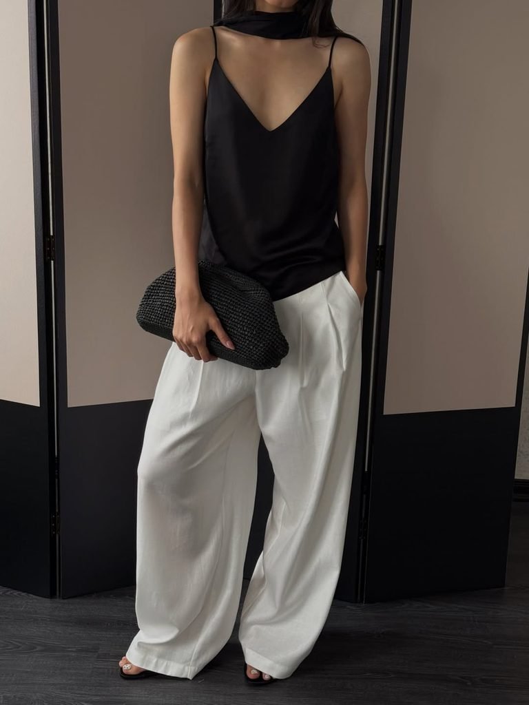 woman wearing black silk camisole and white pleated wide leg trousers holding clutch