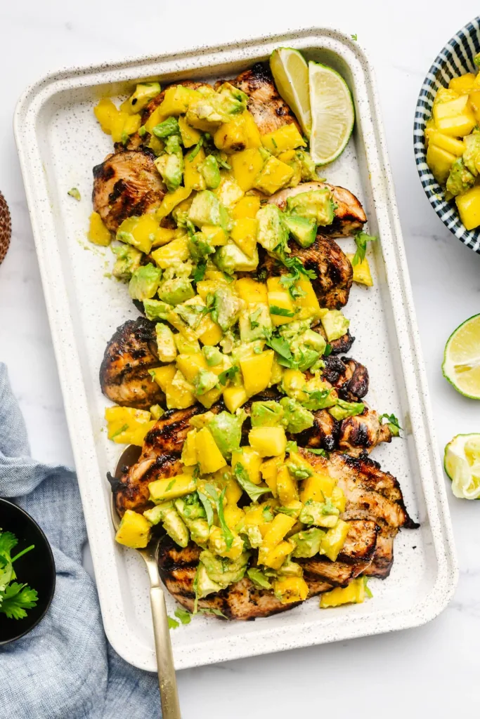 grilled chicken breasts on a serving platter topped with diced mango, avocado, and lime wedges