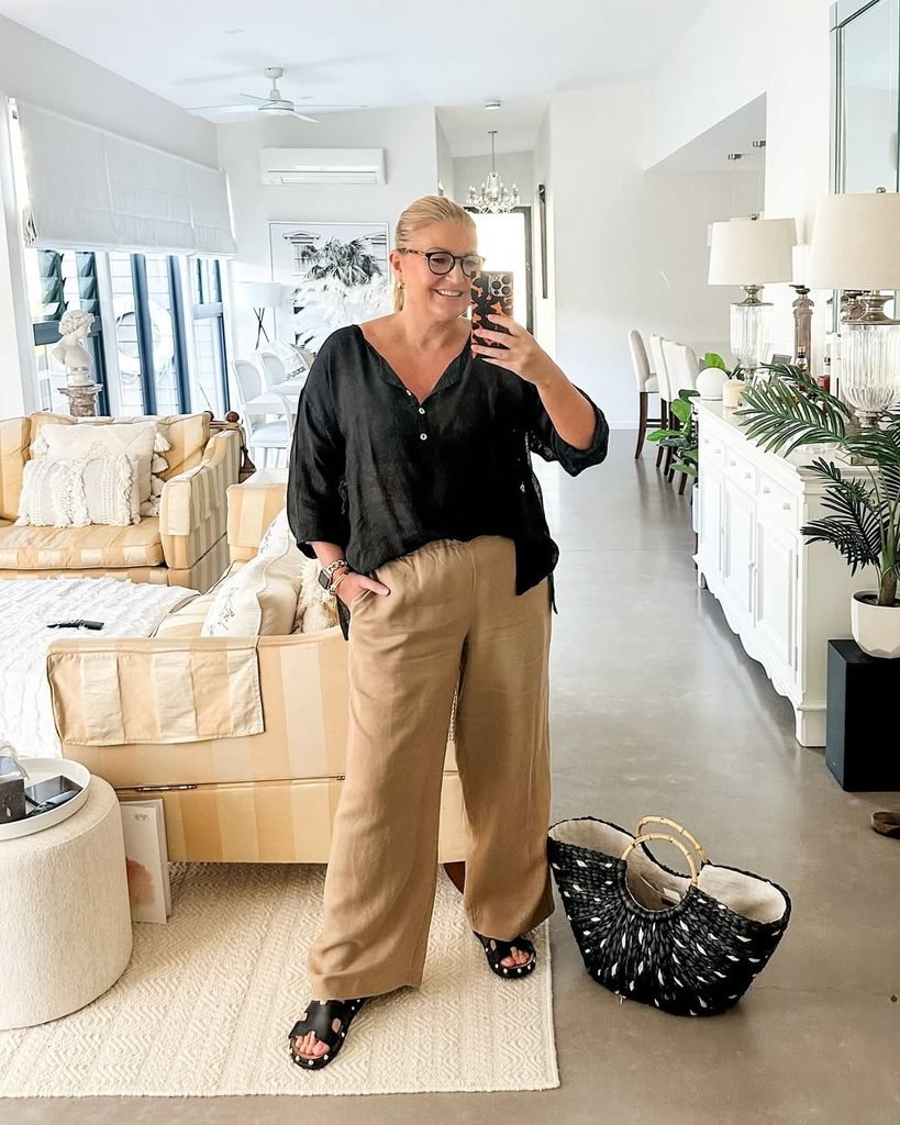 woman in a black top and tan linen pants taking a mirror selfie