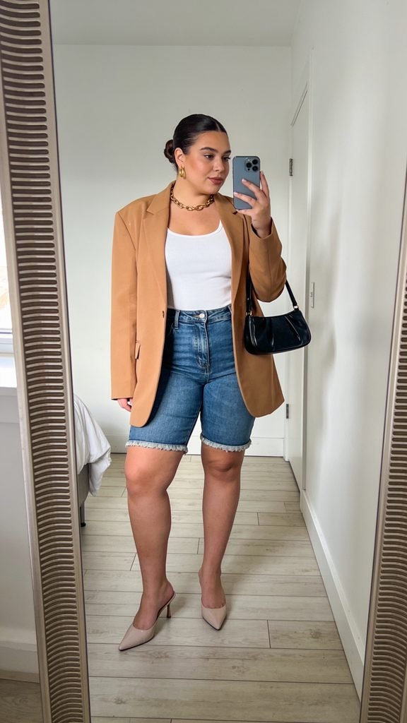 woman in a brown blazer, white top, and denim shorts taking a mirror selfie
