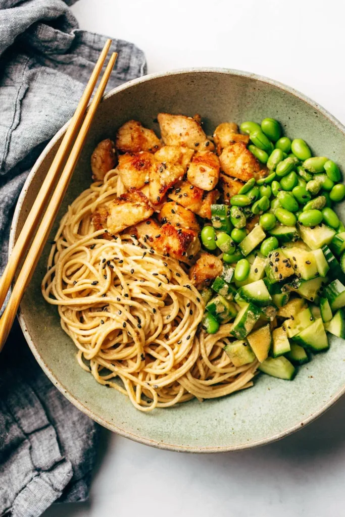 sesame noodles with cooked chicken pieces, edamame, and diced cucumber in a bowl