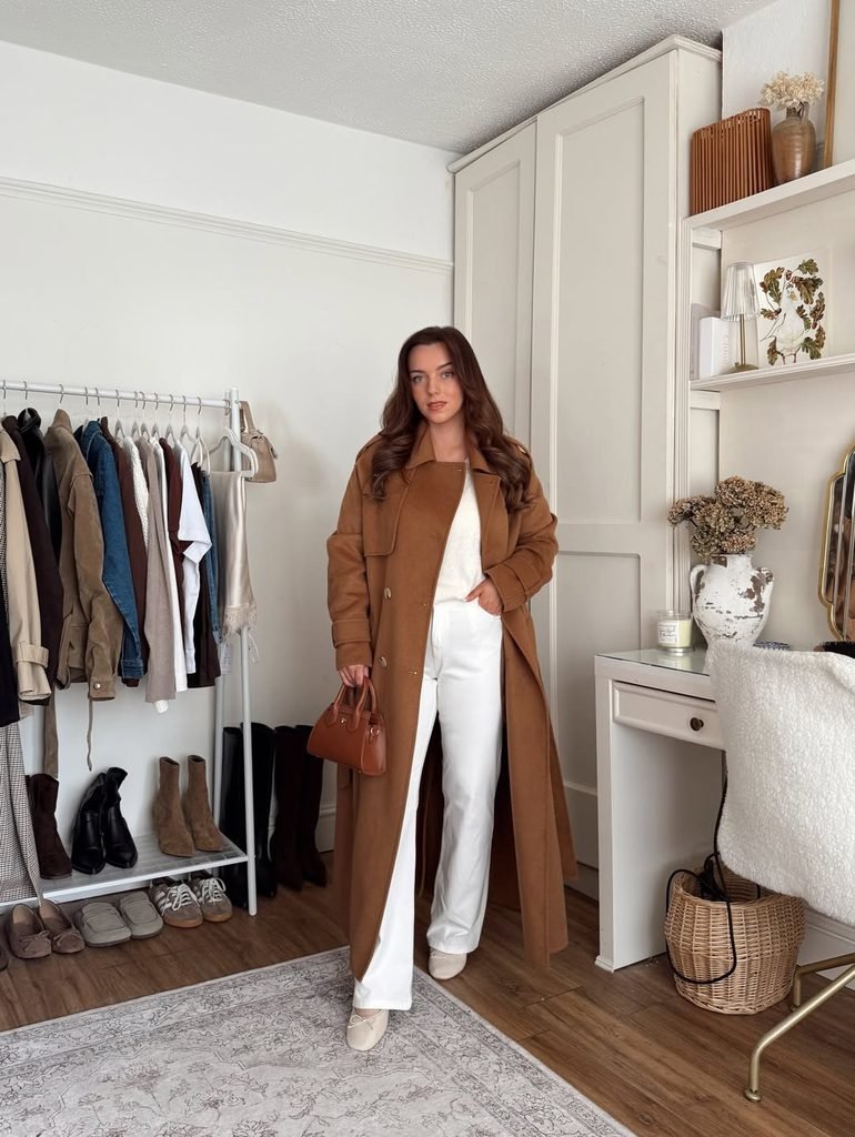 woman in a long brown coat and white jeans carrying a small handbag