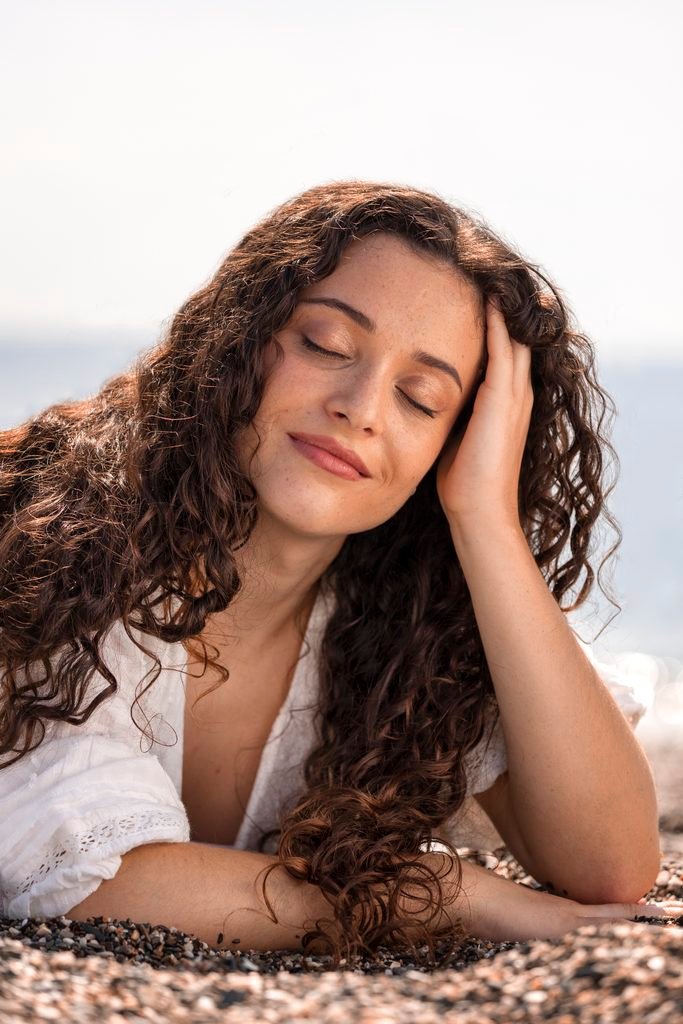 smiling young woman with long curly hair resting on a pebble beach