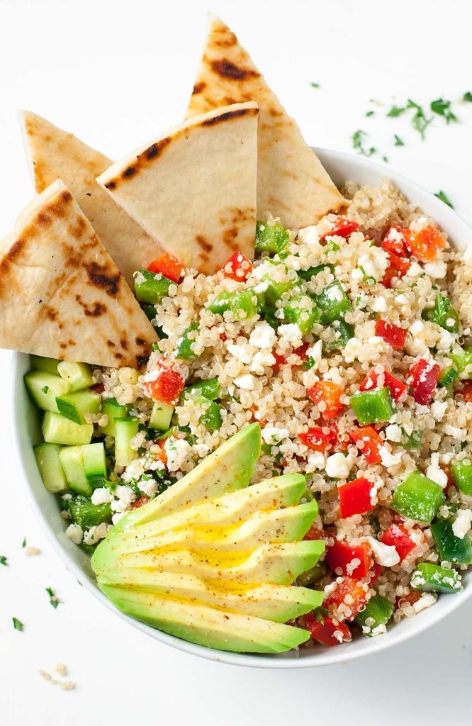 greek quinoa salad bowl topped with sliced avocado, feta cheese, and pita bread wedges