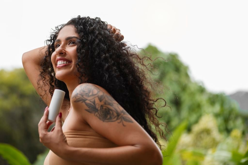 smiling woman with long curly hair holding a small skincare bottle outdoors