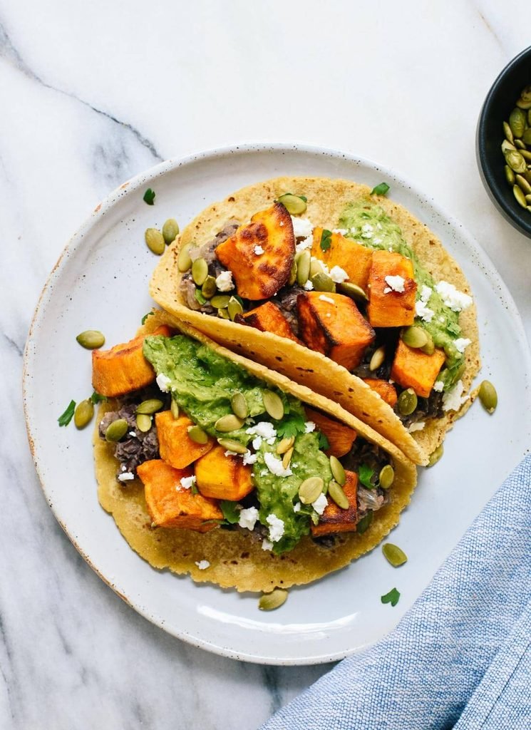 two sweet potato black bean tacos on a plate topped with avocado mash and pepitas
