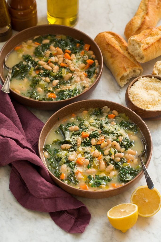 two bowls of white bean kale soup served with sliced baguette and lemon halves