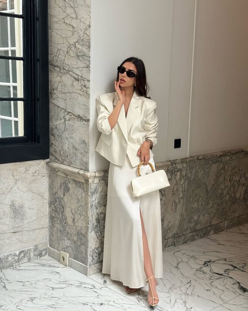 woman wearing a cropped white blazer and long satin skirt leaning against marble wall
