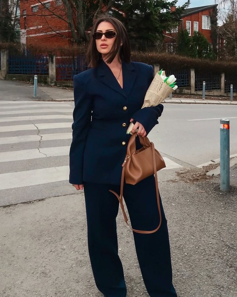 woman in navy pantsuit holding flowers and a brown handbag outdoors