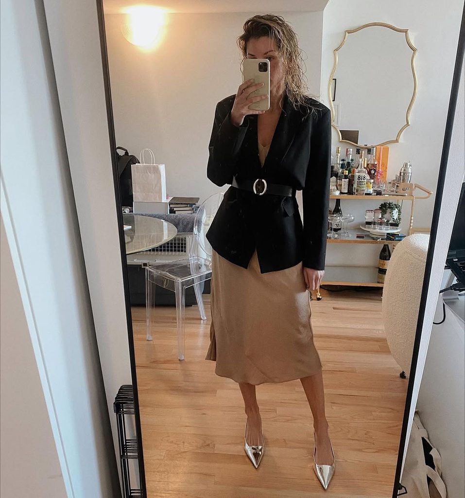 woman in black belted blazer and satin skirt taking a mirror selfie