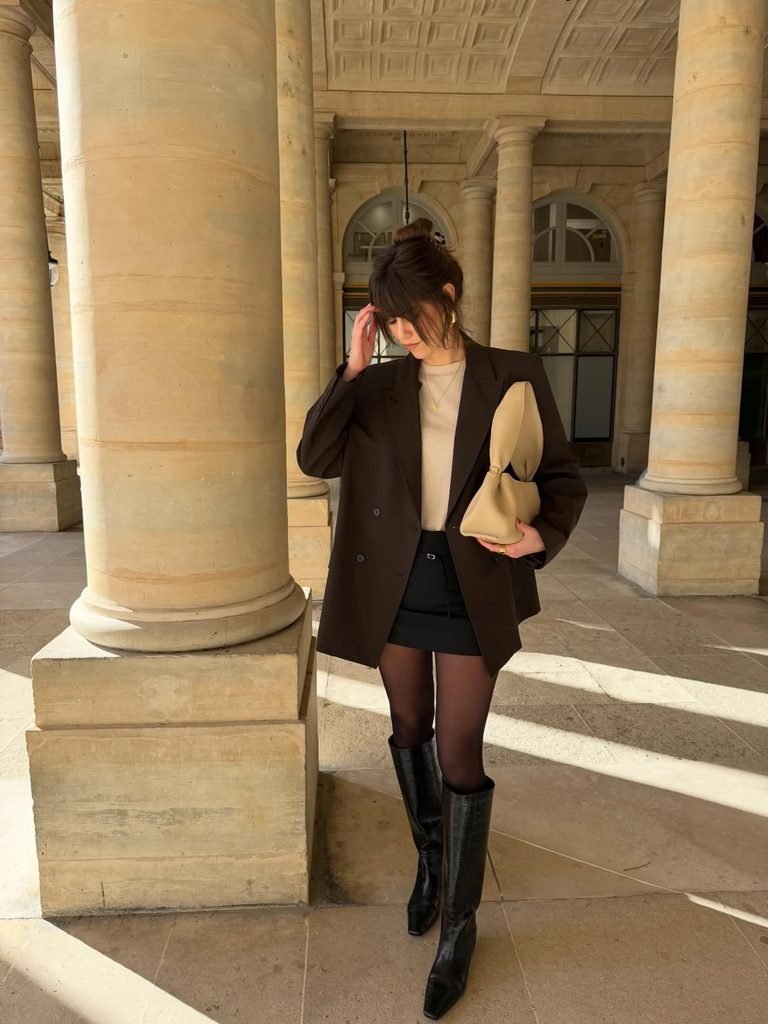 woman standing by columns wearing a brown blazer, black skirt, and boots