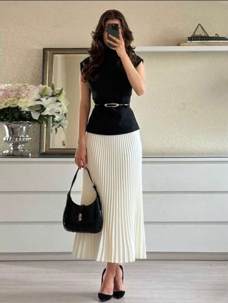 woman wearing a black sleeveless top and white pleated midi skirt holding a handbag