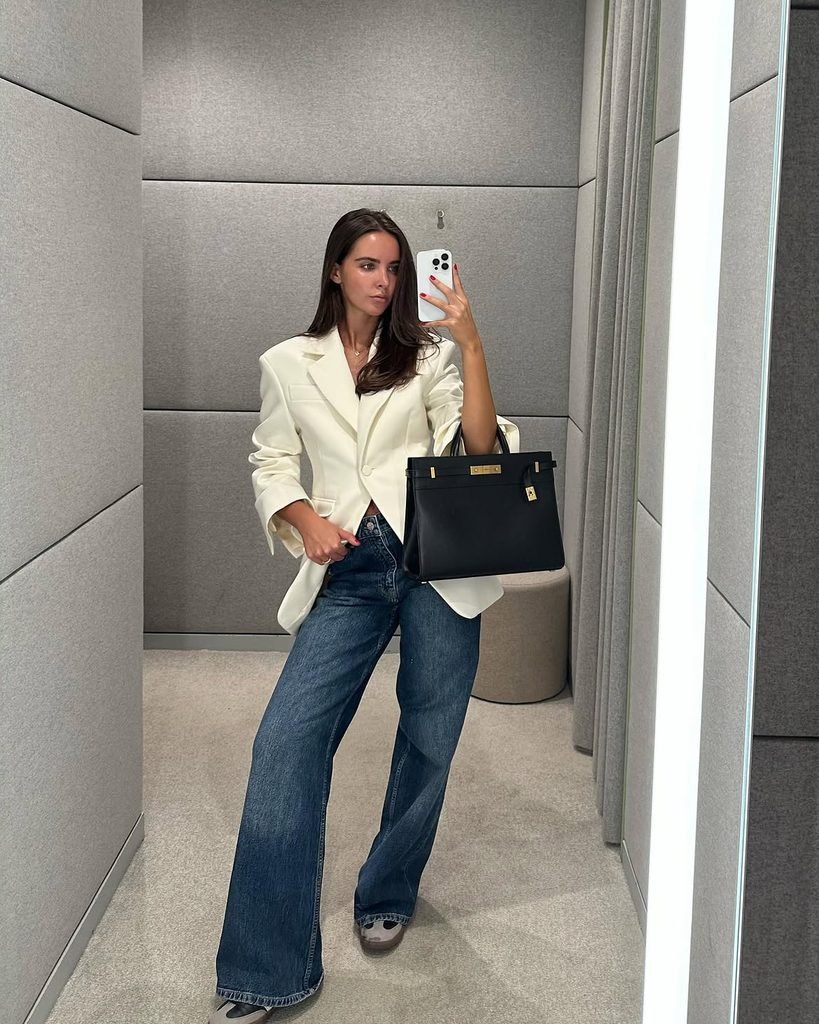 woman taking a mirror selfie wearing a cream blazer blue jeans and black handbag