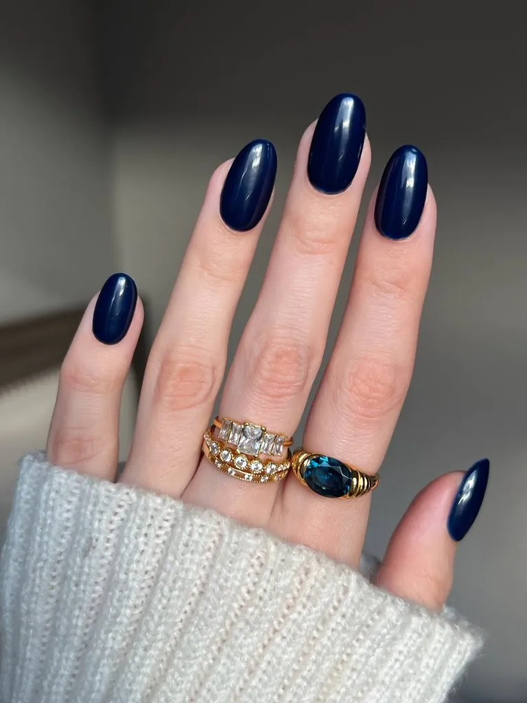 glossy navy blue nails on a hand wearing gold rings and white knit sweater