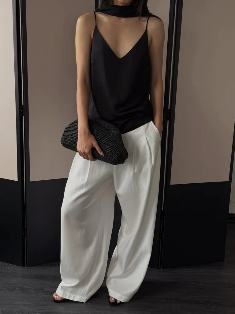 woman wearing black silk camisole and white pleated wide leg trousers holding clutch