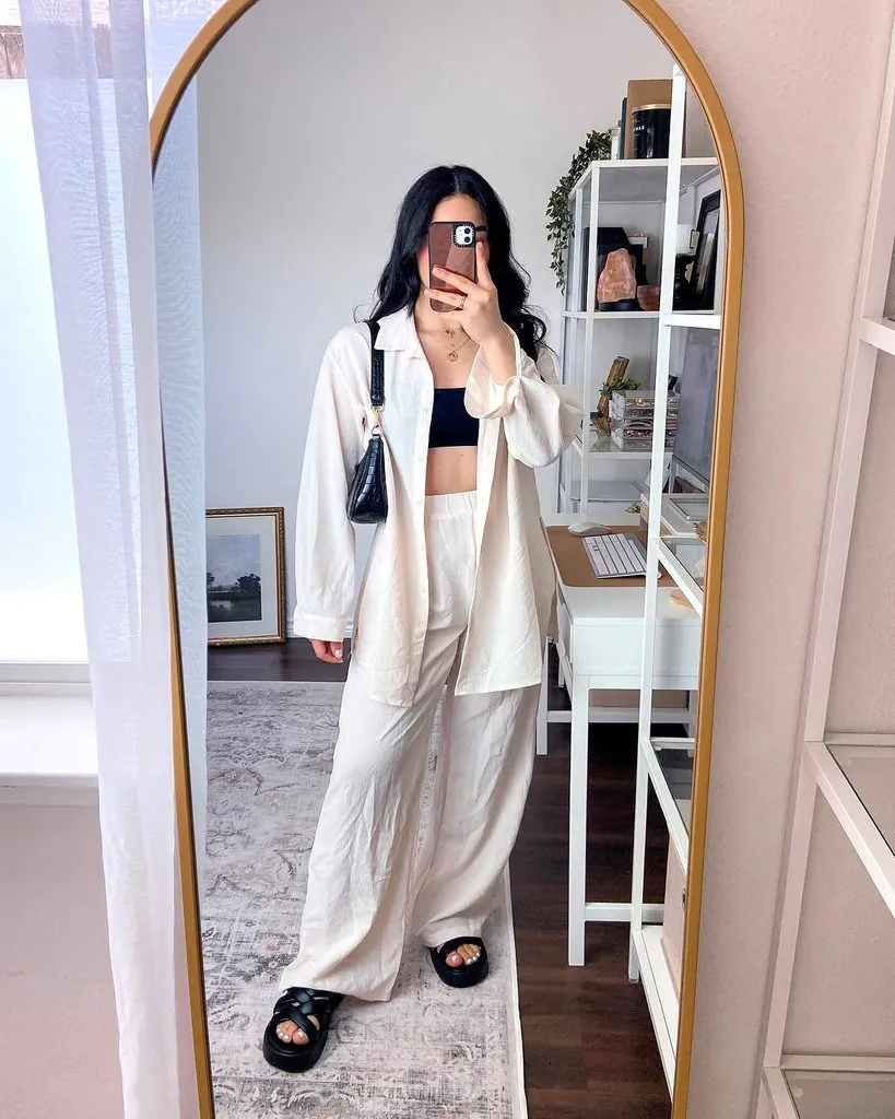 woman taking mirror selfie in cream button down shirt black crop top and trousers