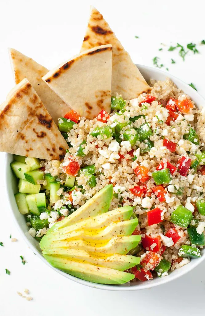 greek quinoa salad bowl topped with sliced avocado, feta cheese, and pita bread wedges