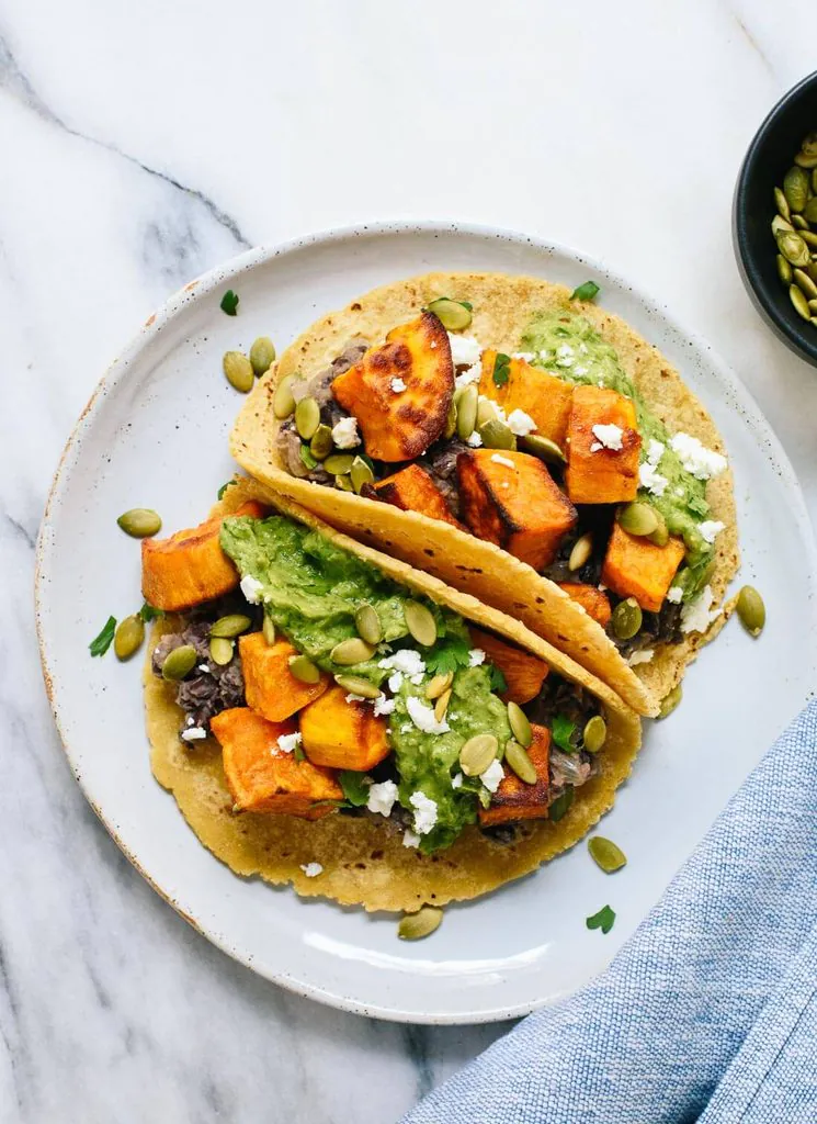 two sweet potato black bean tacos on a plate topped with avocado mash and pepitas