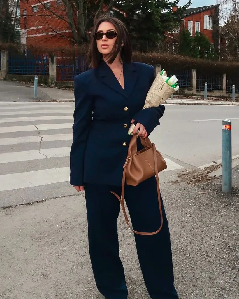 woman in navy pantsuit holding flowers and a brown handbag outdoors