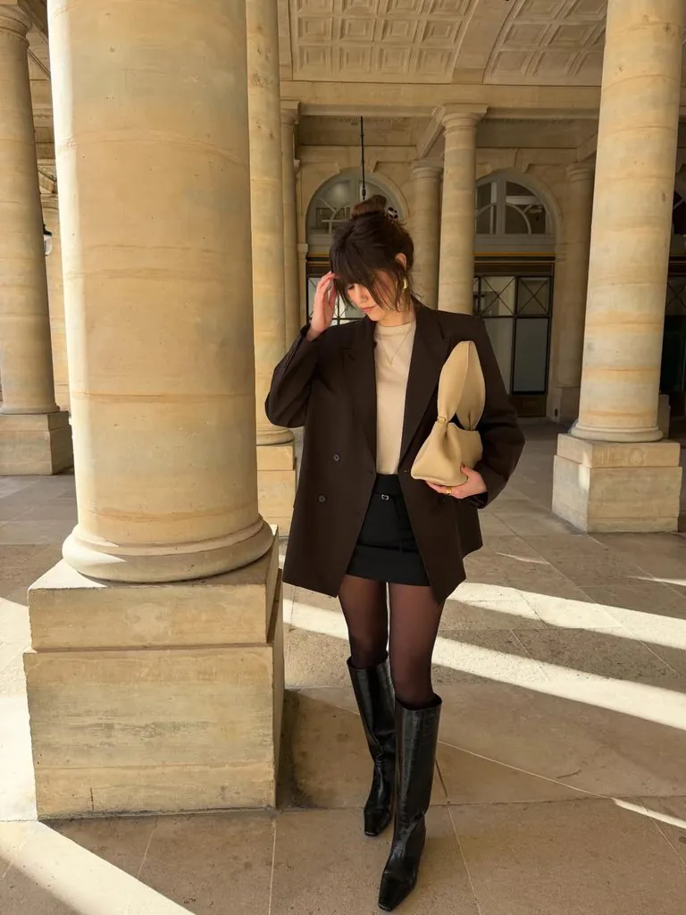 woman standing by columns wearing a brown blazer, black skirt, and boots