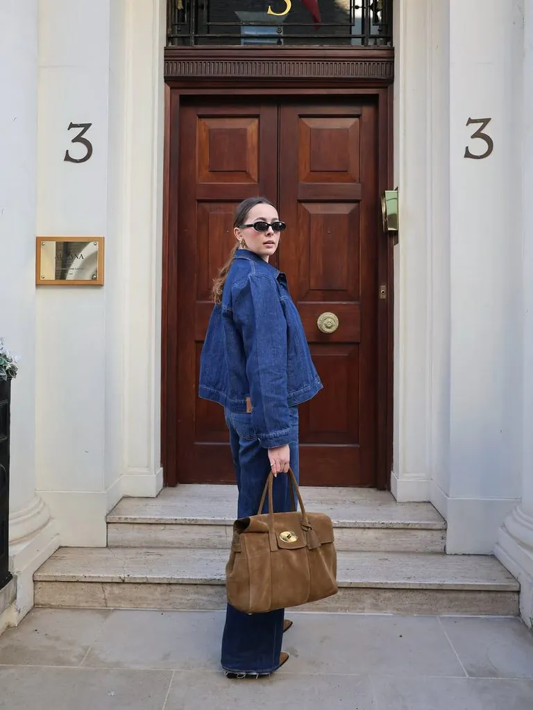 woman in denim jacket and jeans holding a brown suede bag outside