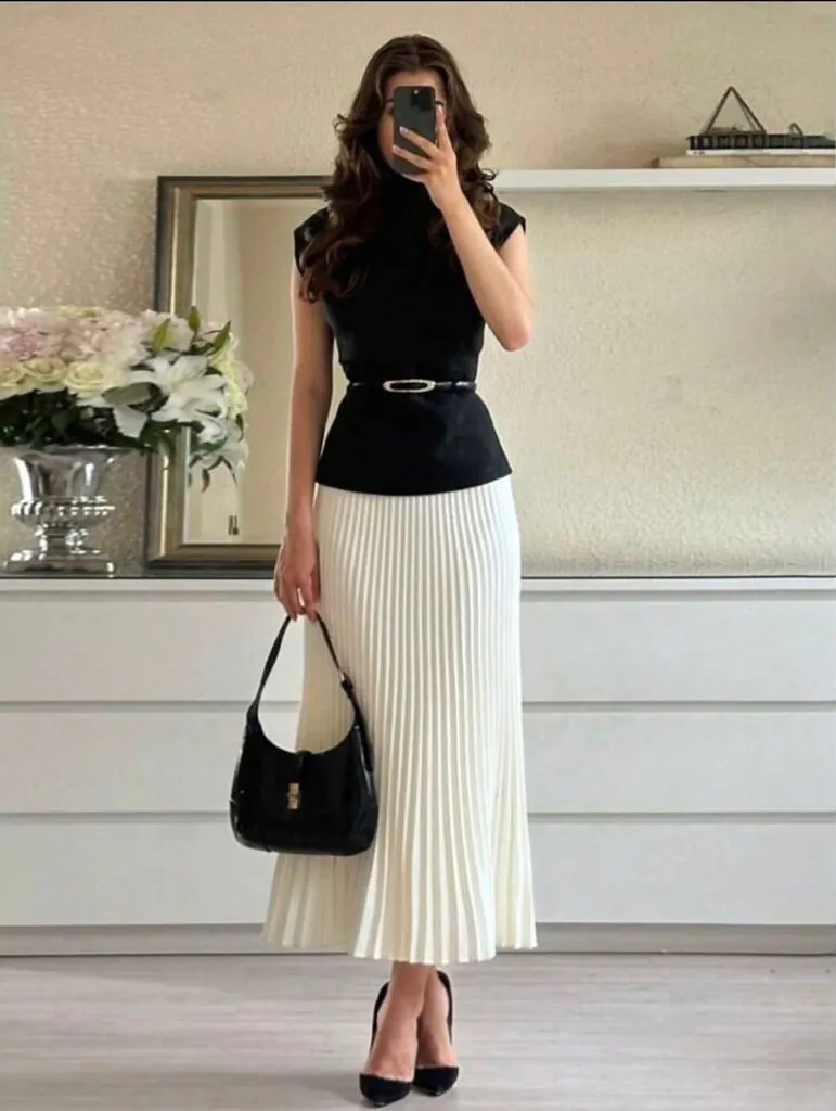 woman wearing a black sleeveless top and white pleated midi skirt holding a handbag