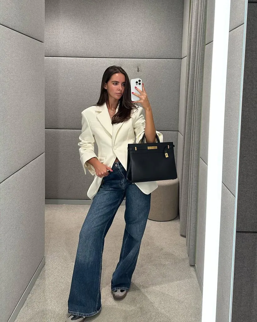 woman taking a mirror selfie wearing a cream blazer blue jeans and black handbag